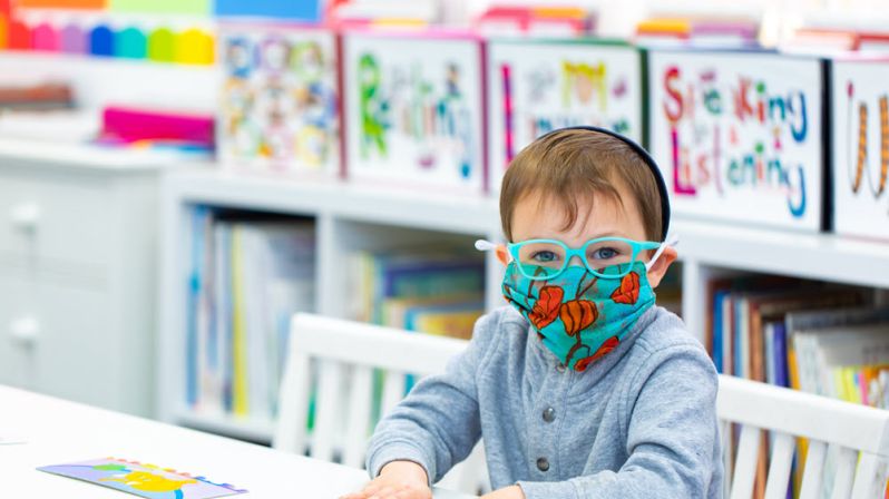Small boy in turquoise color framed glasses and mask reading.